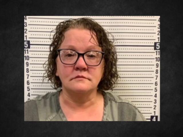 Mugshot of a middle-aged woman with glasses in a gray top, standing before a height-measure backdrop.