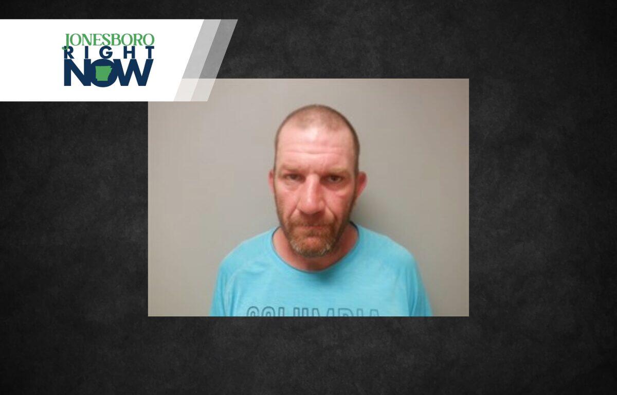 Mugshot-style photo of a middle-aged man with a short beard wearing a light blue shirt, against a gray wall, with Jonesboro Now branding in the corner
