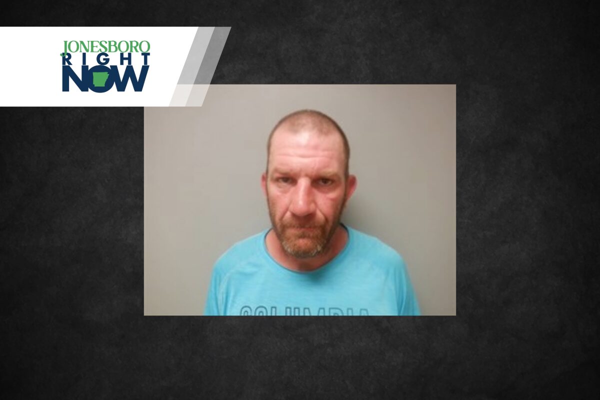 Mugshot-style photo of a middle-aged man with a short beard wearing a light blue shirt, against a gray wall, with Jonesboro Now branding in the corner