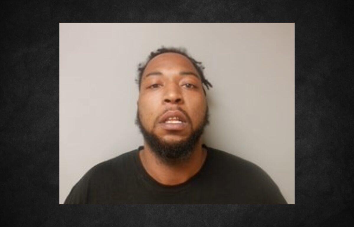 Mugshot of a man with a beard and short dreadlocks wearing a dark shirt, against a light gray background.