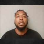 Mugshot of a man with a beard and short dreadlocks wearing a dark shirt, against a light gray background.