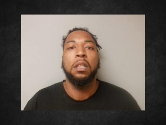 Mugshot of a man with a beard and short dreadlocks wearing a dark shirt, against a light gray background.