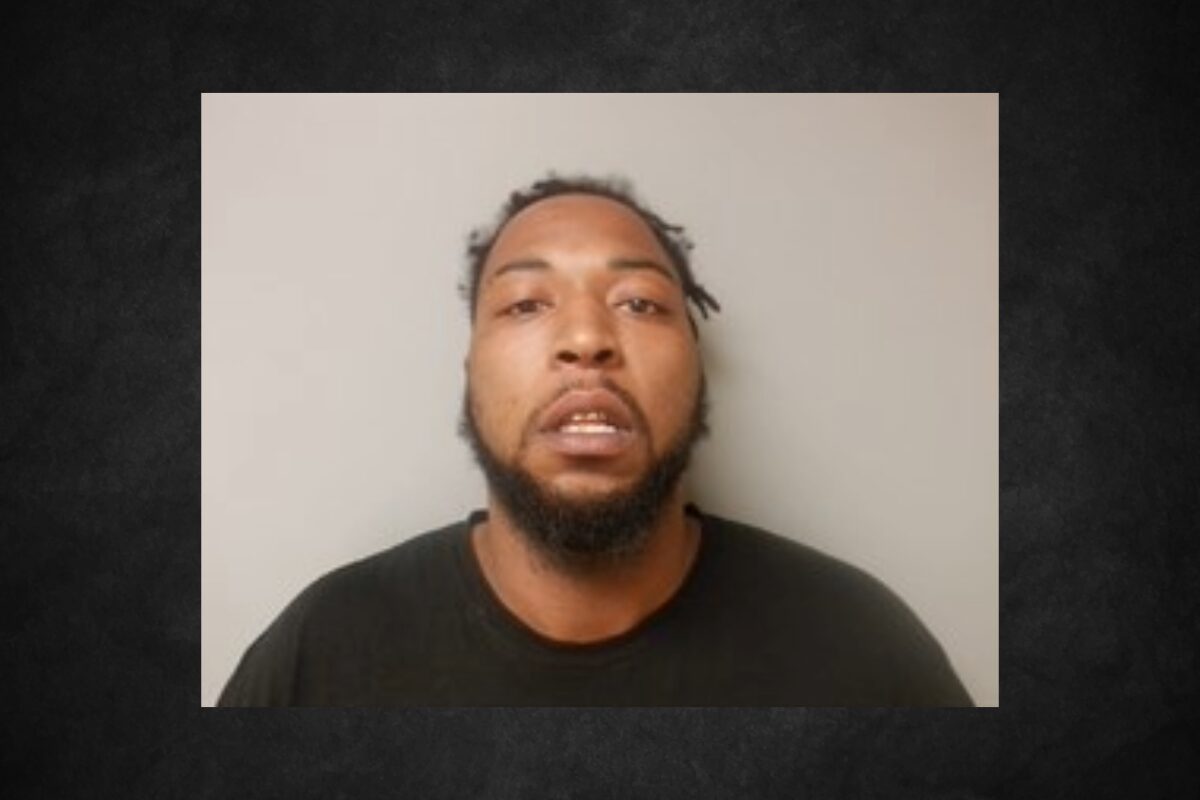 Mugshot of a man with a beard and short dreadlocks wearing a dark shirt, against a light gray background.