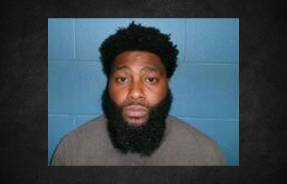 Mugshot of a man with a thick black beard and short curly hair, wearing a tan shirt, against a blue wall backdrop.