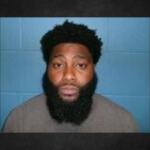 Mugshot of a man with a thick black beard and short curly hair, wearing a tan shirt, against a blue wall backdrop.