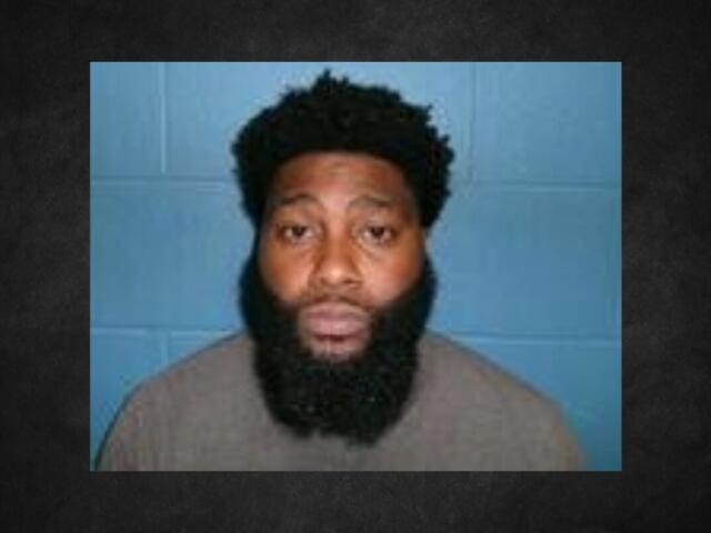 Mugshot of a man with a thick black beard and short curly hair, wearing a tan shirt, against a blue wall backdrop.