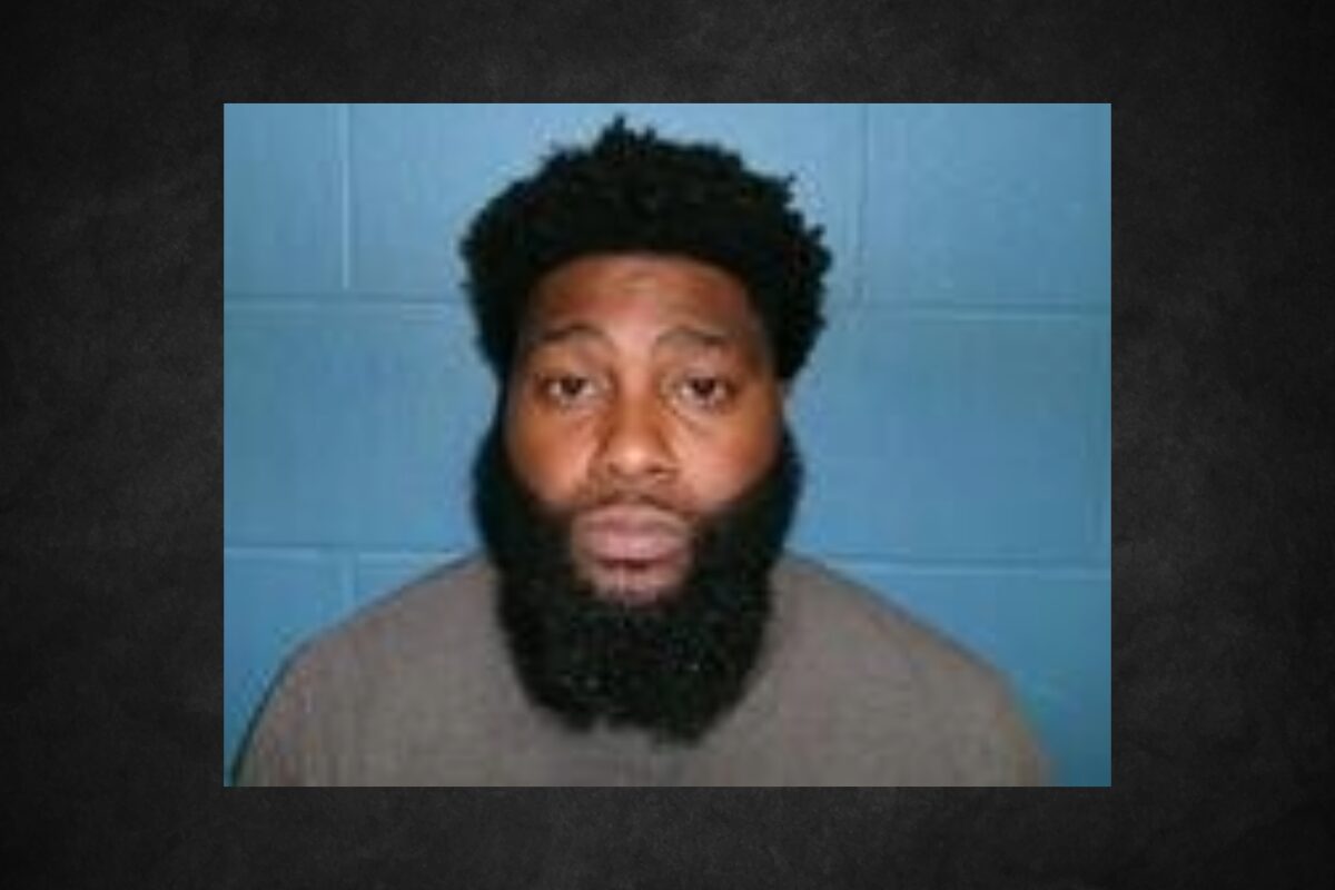 Mugshot of a man with a thick black beard and short curly hair, wearing a tan shirt, against a blue wall backdrop.