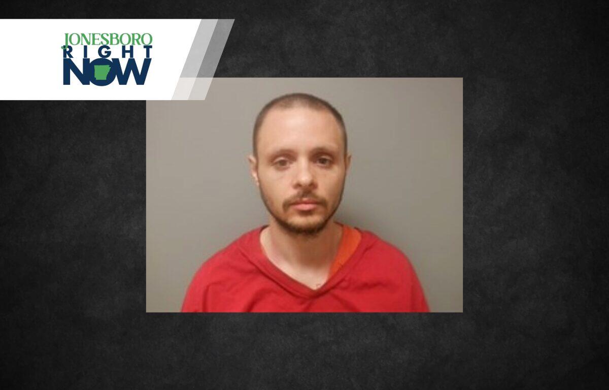 Mugshot-style photo of a man with short hair and beard wearing a red shirt against a gray wall, Jonesboro Now logo in the corner.