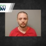 Mugshot-style photo of a man with short hair and beard wearing a red shirt against a gray wall, Jonesboro Now logo in the corner.