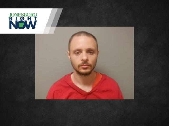 Mugshot-style photo of a man with short hair and beard wearing a red shirt against a gray wall, Jonesboro Now logo in the corner.