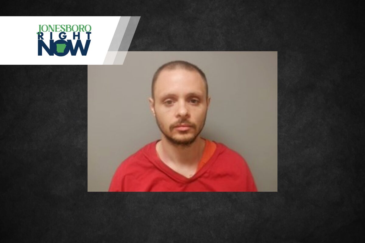 Mugshot-style photo of a man with short hair and beard wearing a red shirt against a gray wall, Jonesboro Now logo in the corner.