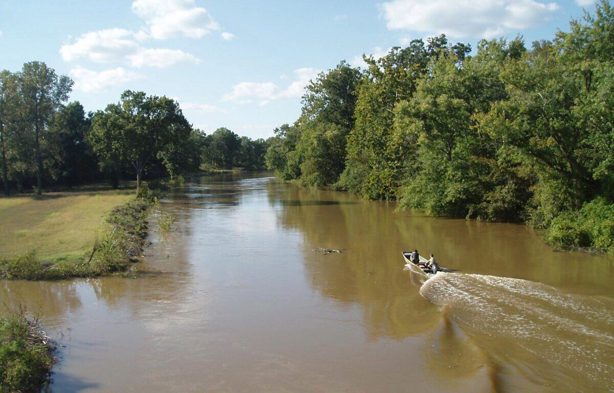 A small motorboat with two people travels along a muddy river bordered by trees under a blue sky.
