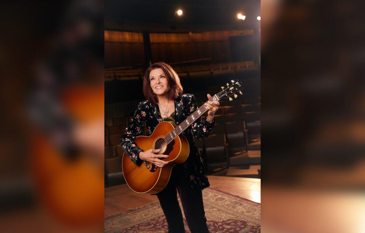 Female musician holding an acoustic guitar on a stage, smiling backstage with warm lighting around her.