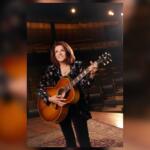 Female musician holding an acoustic guitar on a stage, smiling backstage with warm lighting around her.