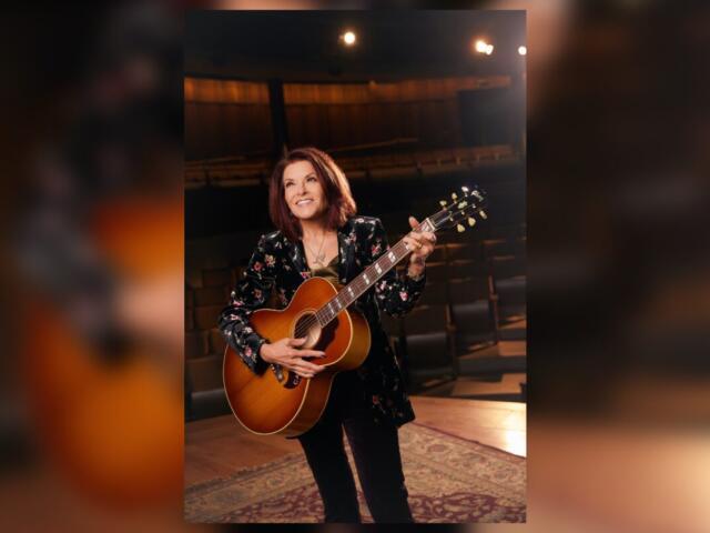Female musician holding an acoustic guitar on a stage, smiling backstage with warm lighting around her.