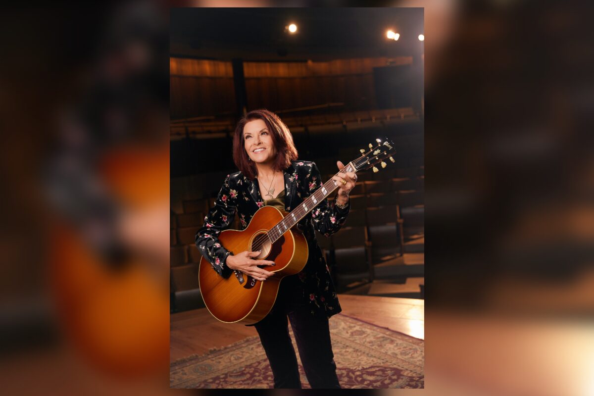 Female musician holding an acoustic guitar on a stage, smiling backstage with warm lighting around her.