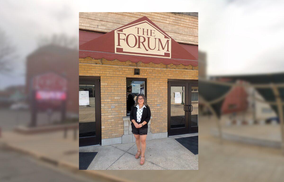 Woman in a black blazer, white blouse, and shorts stands outside The Forum entrance under a maroon awning.