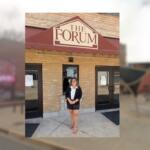 Woman in a black blazer, white blouse, and shorts stands outside The Forum entrance under a maroon awning.