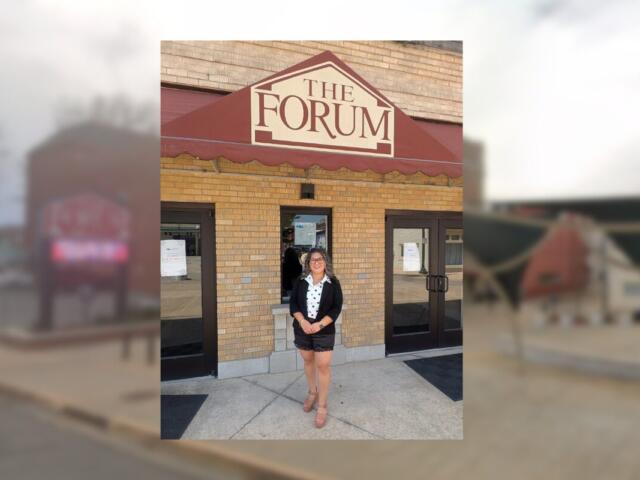 Woman in a black blazer, white blouse, and shorts stands outside The Forum entrance under a maroon awning.