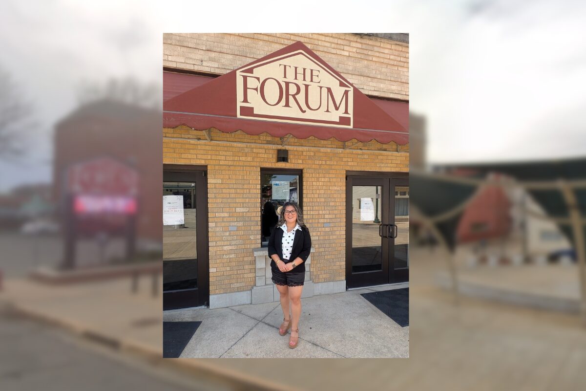 Woman in a black blazer, white blouse, and shorts stands outside The Forum entrance under a maroon awning.