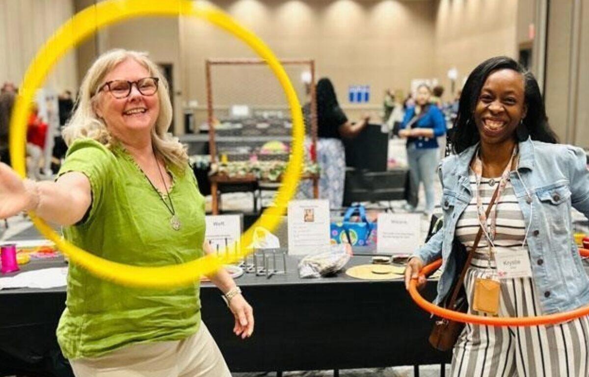 Two women at an indoor booth, one in a green shirt spinning a yellow hoop while the other holds an orange hoop and smiles conference-style event atmosphere.