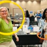Two women at an indoor booth, one in a green shirt spinning a yellow hoop while the other holds an orange hoop and smiles conference-style event atmosphere.