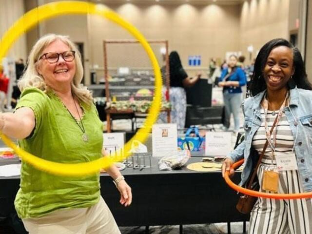 Two women at an indoor booth, one in a green shirt spinning a yellow hoop while the other holds an orange hoop and smiles conference-style event atmosphere.