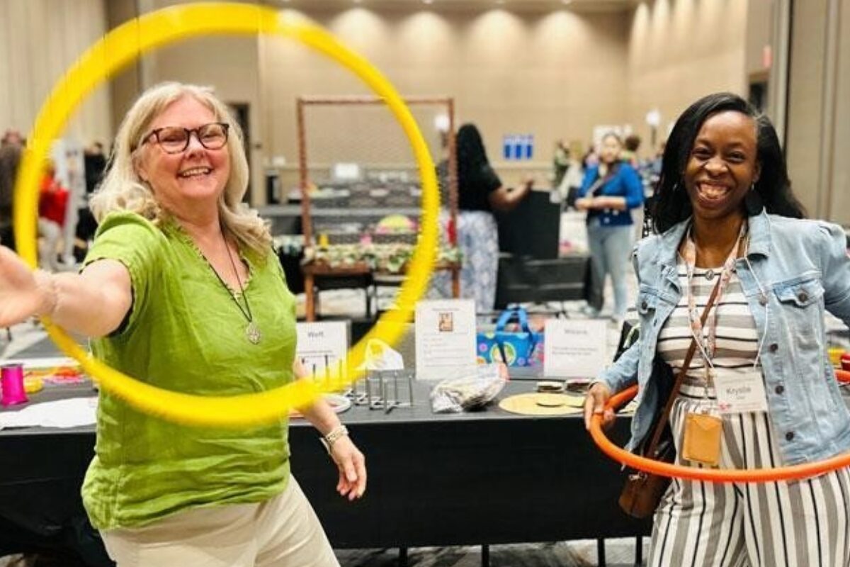 Two women at an indoor booth, one in a green shirt spinning a yellow hoop while the other holds an orange hoop and smiles conference-style event atmosphere.