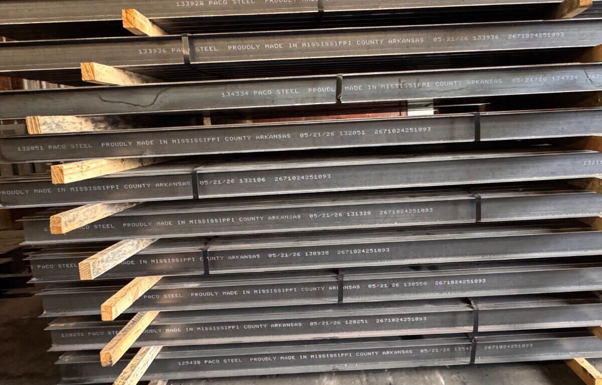 Stacked steel beams with white stamped lettering and wooden spacers in a warehouse.