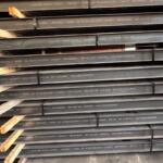 Stacked steel beams with white stamped lettering and wooden spacers in a warehouse.