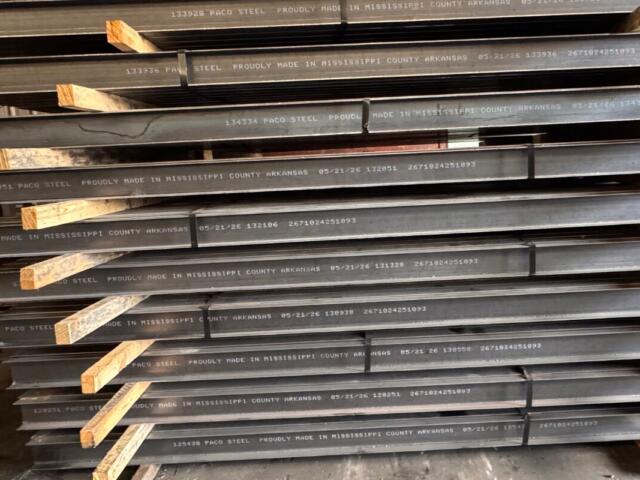 Stacked steel beams with white stamped lettering and wooden spacers in a warehouse.