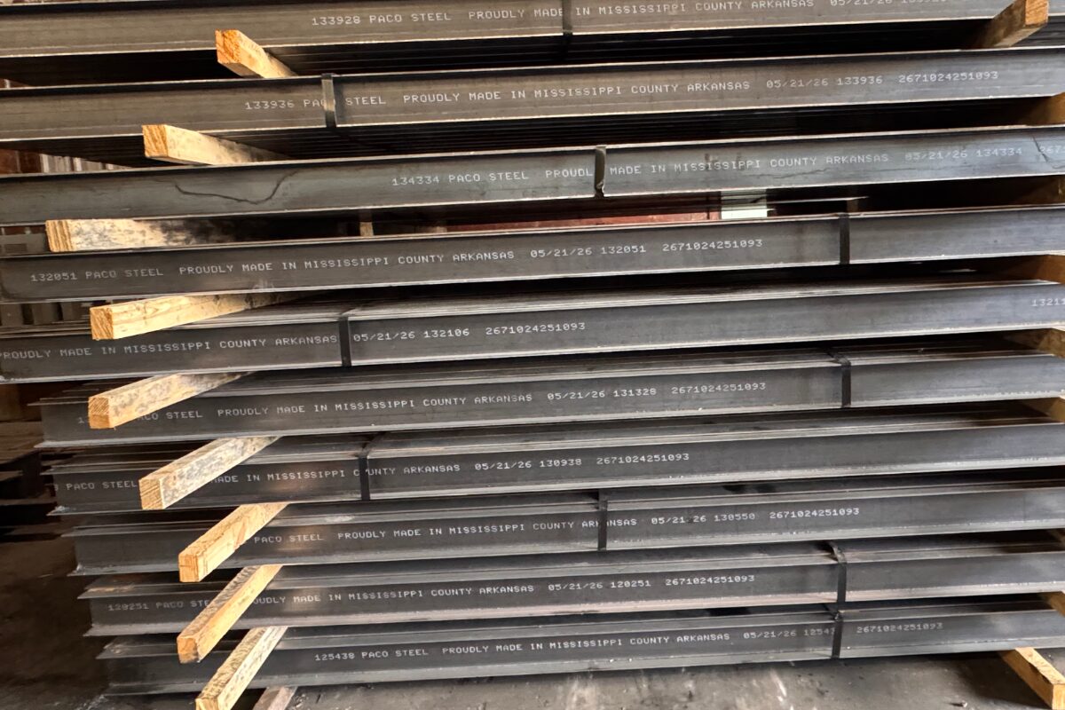 Stacked steel beams with white stamped lettering and wooden spacers in a warehouse.