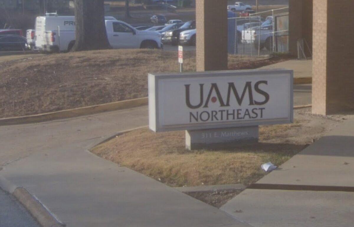 Sign reading 'UAMS Northeast' mounted on a low pedestal in a landscaped area near a walkway, with a parking lot and vehicles in the background.