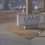 Sign reading 'UAMS Northeast' mounted on a low pedestal in a landscaped area near a walkway, with a parking lot and vehicles in the background.