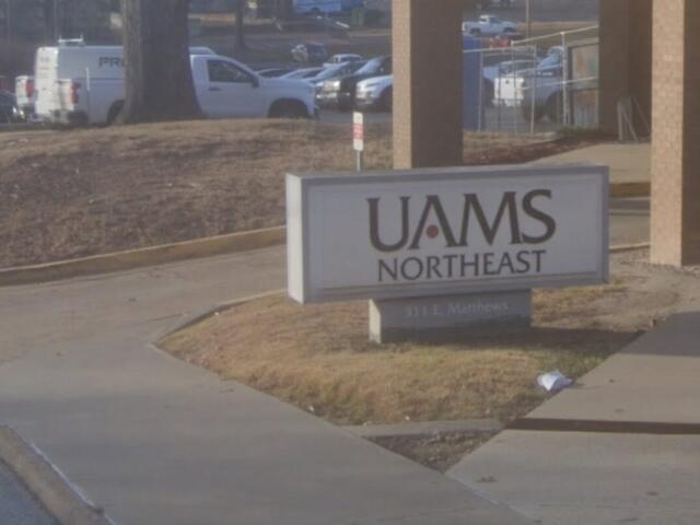 Sign reading 'UAMS Northeast' mounted on a low pedestal in a landscaped area near a walkway, with a parking lot and vehicles in the background.