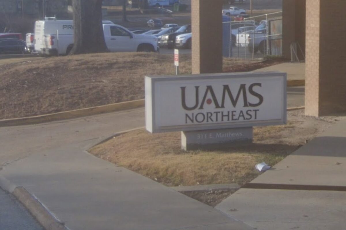 Sign reading 'UAMS Northeast' mounted on a low pedestal in a landscaped area near a walkway, with a parking lot and vehicles in the background.