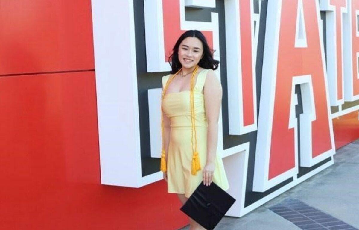Smiling woman in a yellow dress poses with a black clutch in front of large red and white 3D letters on an outdoor wall near her.
