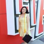 Smiling woman in a yellow dress poses with a black clutch in front of large red and white 3D letters on an outdoor wall near her.