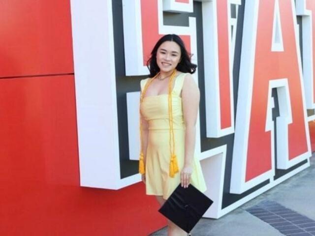 Smiling woman in a yellow dress poses with a black clutch in front of large red and white 3D letters on an outdoor wall near her.