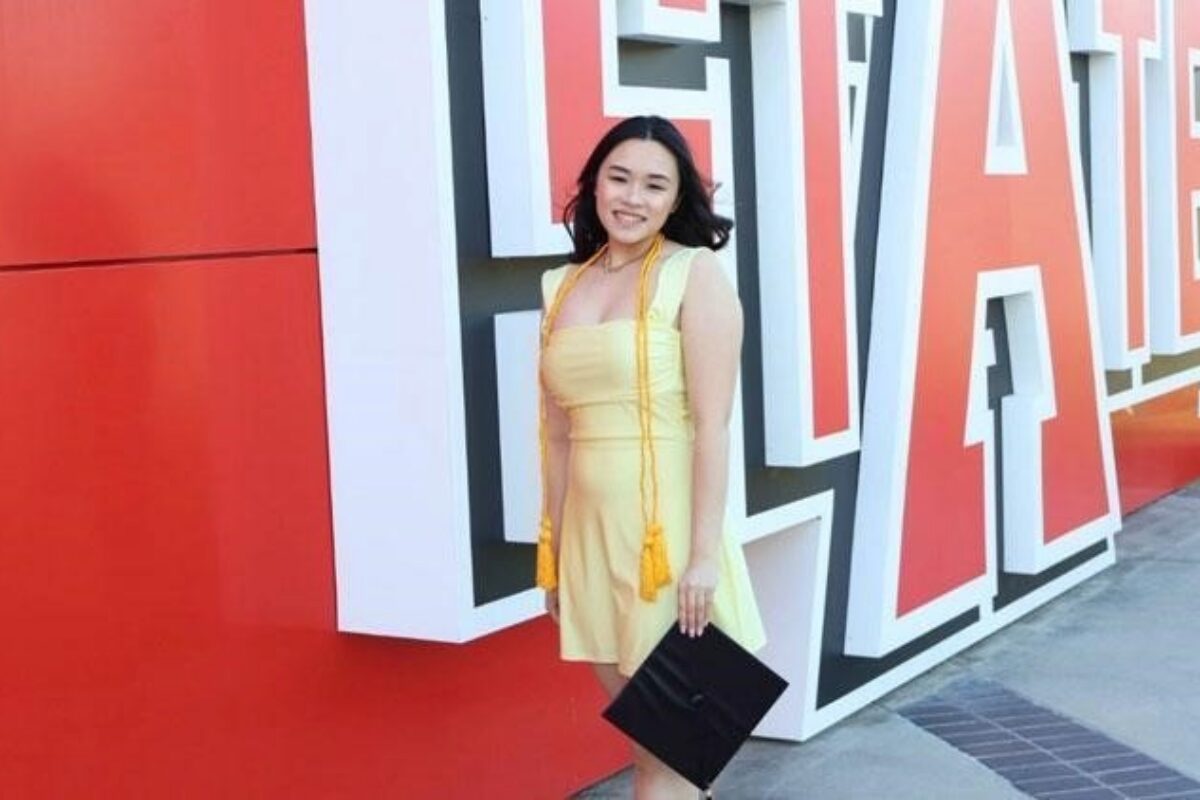 Smiling woman in a yellow dress poses with a black clutch in front of large red and white 3D letters on an outdoor wall near her.