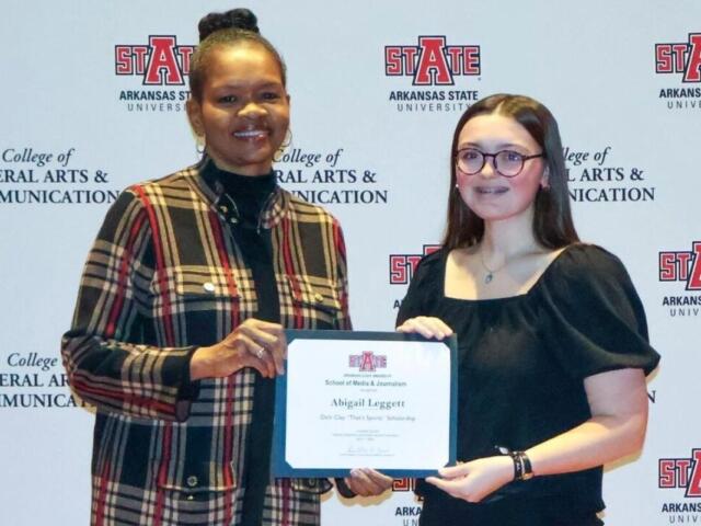 Two women pose with a scholarship certificate at an Arkansas State University backdrop (College of Liberal Arts & Communications).