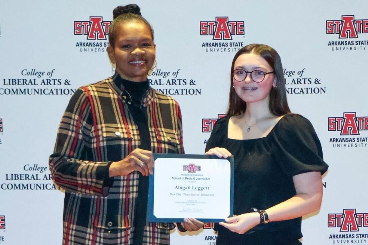 Two women pose with a scholarship certificate at an Arkansas State University backdrop (College of Liberal Arts & Communications).