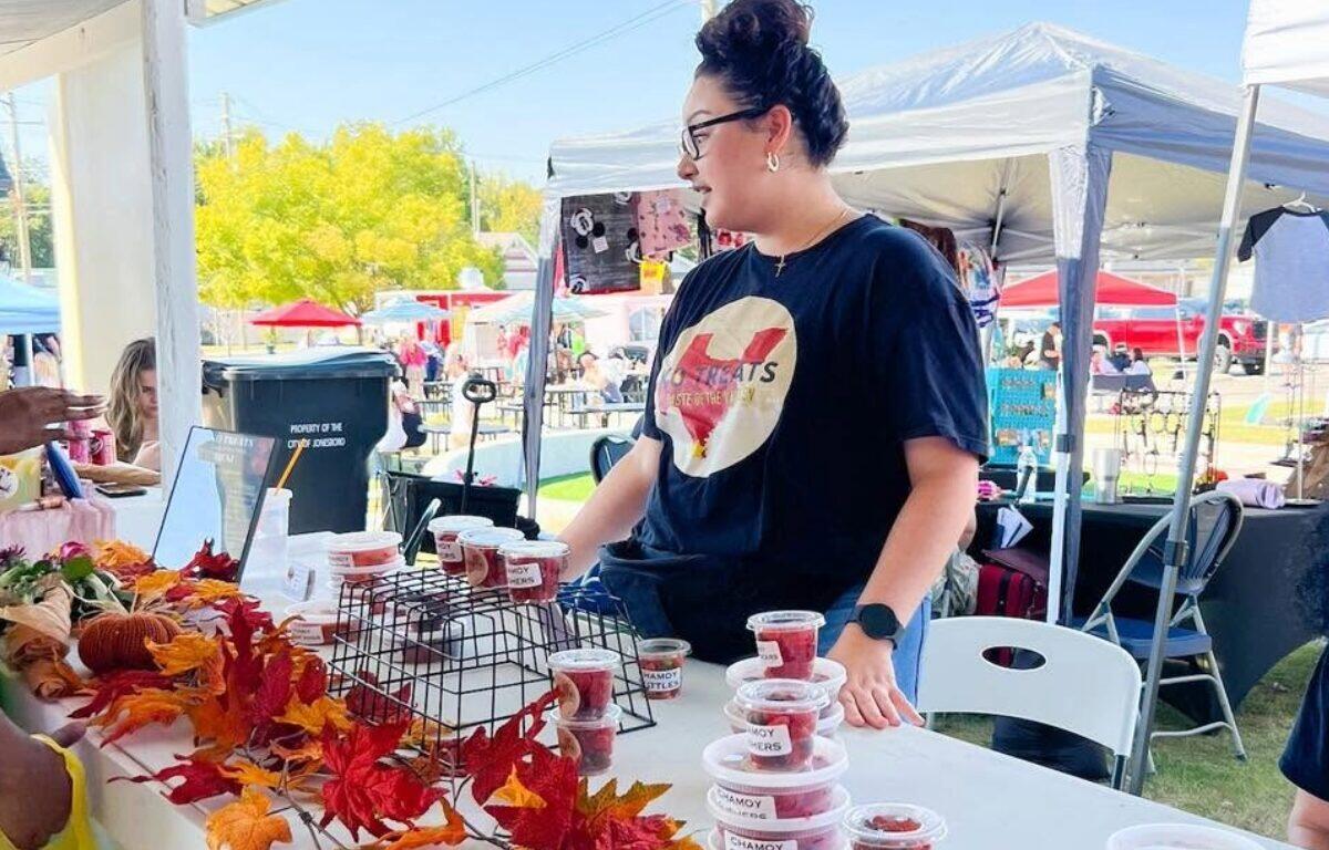 Vendor at an outdoor market stall with cups of pink chamoy snacks, autumn leaf decor on the table, and a shopper nearby.