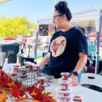 Vendor at an outdoor market stall with cups of pink chamoy snacks, autumn leaf decor on the table, and a shopper nearby.