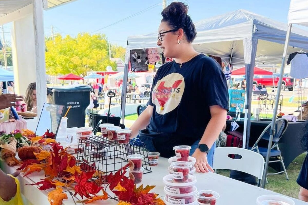 Vendor at an outdoor market stall with cups of pink chamoy snacks, autumn leaf decor on the table, and a shopper nearby.