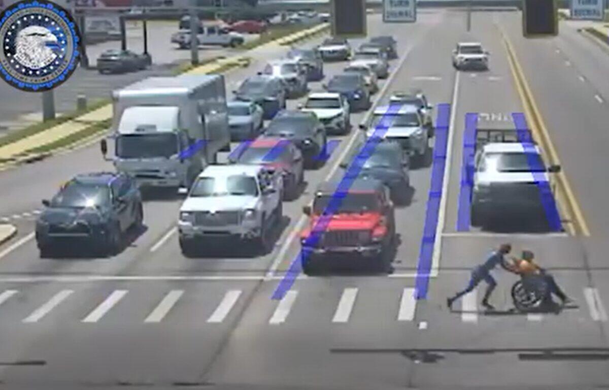 Two people push a person in a wheelchair across a crosswalk at a busy multi-lane intersection as cars wait in traffic, with blue lane guidance markings visible on the road.