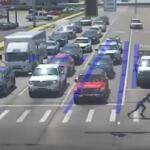 Two people push a person in a wheelchair across a crosswalk at a busy multi-lane intersection as cars wait in traffic, with blue lane guidance markings visible on the road.
