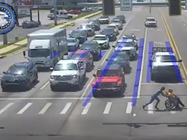 Two people push a person in a wheelchair across a crosswalk at a busy multi-lane intersection as cars wait in traffic, with blue lane guidance markings visible on the road.
