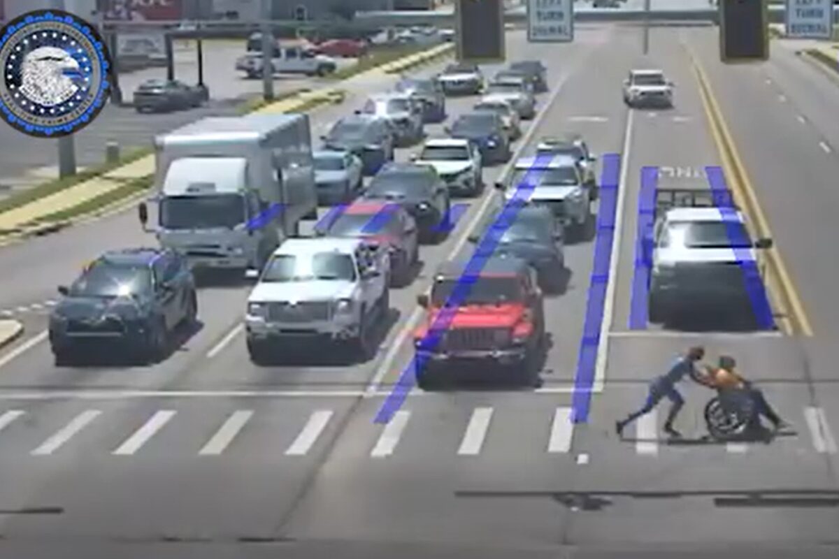 Two people push a person in a wheelchair across a crosswalk at a busy multi-lane intersection as cars wait in traffic, with blue lane guidance markings visible on the road.