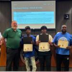 Six people stand behind a council desk; four award recipients hold certificates, flanked by officials during City Council meeting.